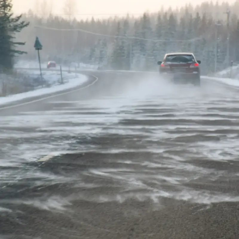 A red car driving on a snowy highway as wind blows snow across the icy road between forested shoulders.
