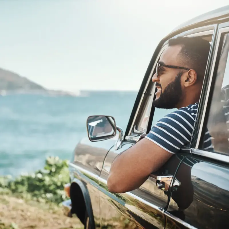 A man in sunglasses smiles with his arm resting on the open window of a car parked beside the ocean on a sunny day.