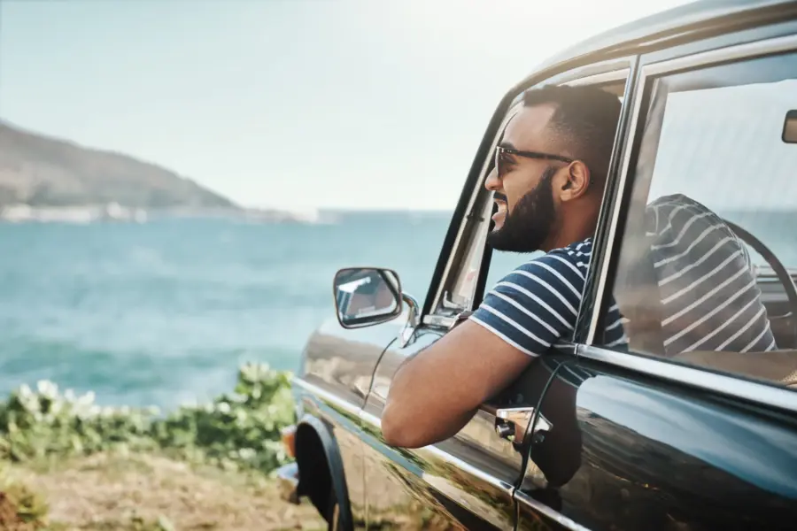 A man in sunglasses smiles with his arm resting on the open window of a car parked beside the ocean on a sunny day.