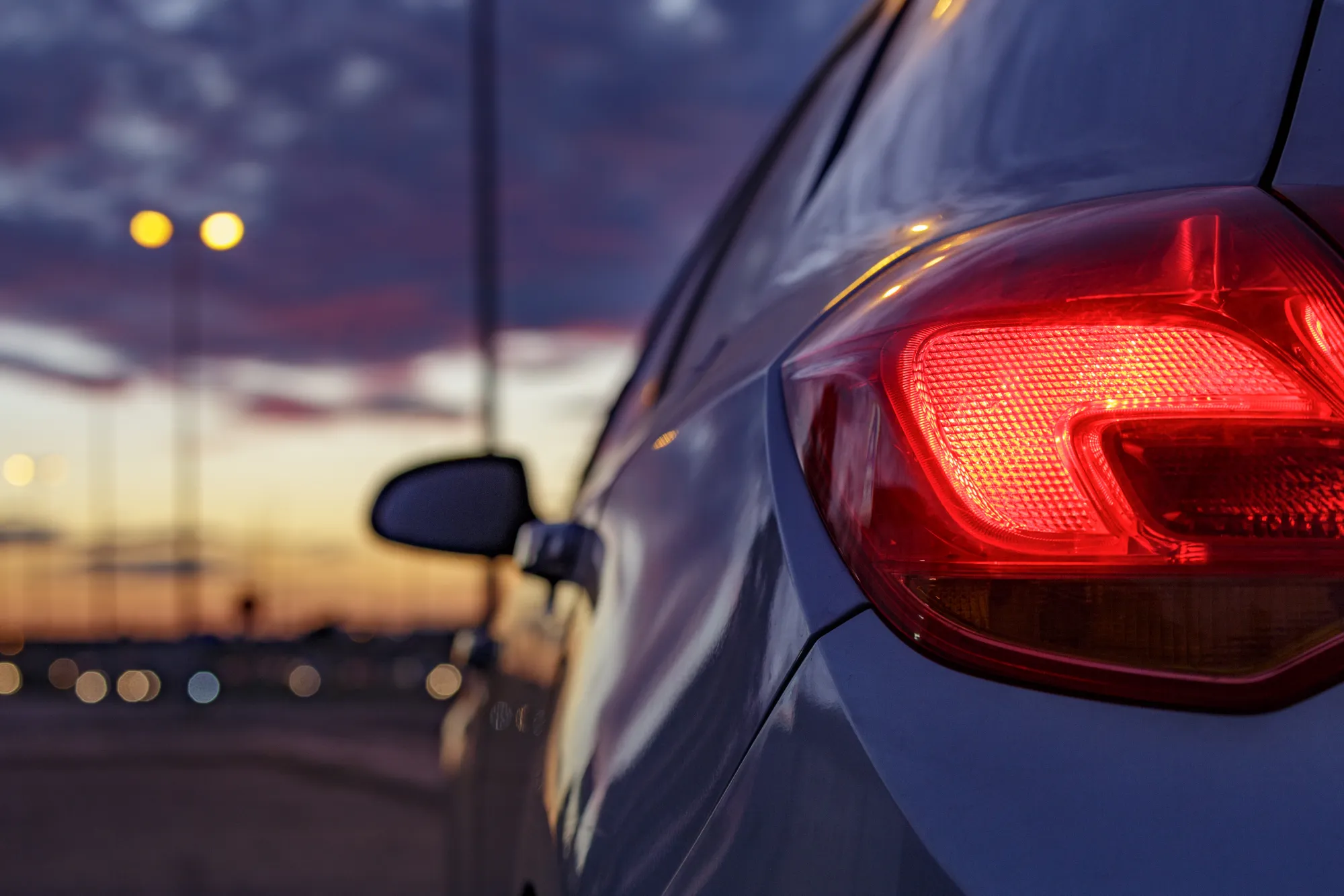 Illuminated red taillight on the rear corner of a dark blue car parked beside a road at dusk.